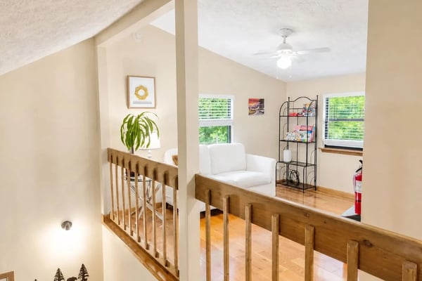 Loft bedroom with wooden railing, white armchair, potted plant, and windows overlooking greenery